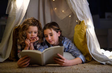 Sisters indoors at home, reading book in pajamas in tent. Lockdown concept. - HPIF05740