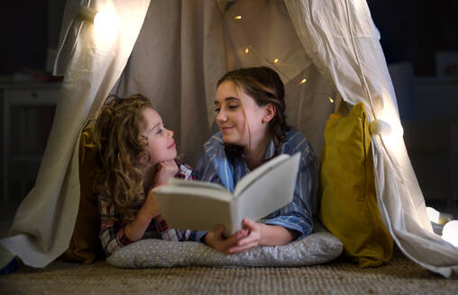 Sisters indoors at home, reading book in pajamas in tent. Lockdown concept. - HPIF05741