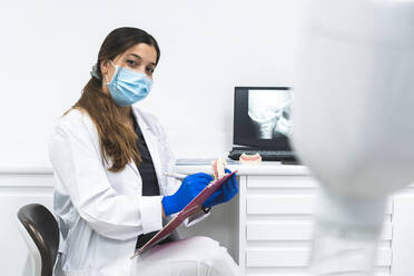 Young dentist wearing surgical mask sitting with clipboard in clinic - JAQF01190