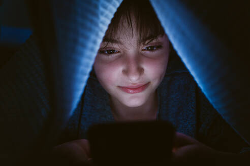 Ein glückliches Teenager-Mädchen, das sein Smartphone benutzt und sich nachts unter einer Decke versteckt, um soziale Netzwerke zu nutzen. - HPIF05787