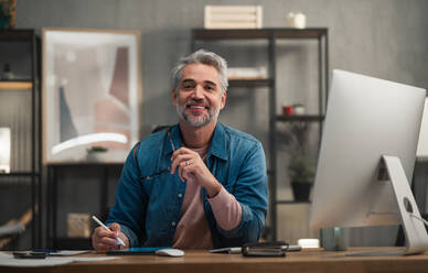 A happy mature man architect working on tablet at desk indoors in office, looking at camera. - HPIF05803