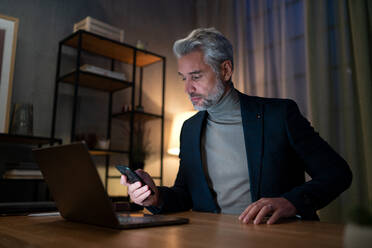 A mature businessman working on laptop at desk indoors in office at night. - HPIF05829