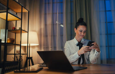 A happy mid adult business woman with diary working on laptop in office at night, looking at camera. - HPIF05832