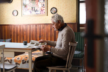 A portrait of elderly man sitting at the table indoors at home, reading newspapers. - HPIF05865