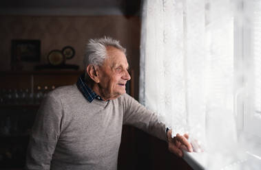 A portrait of elderly man standing indoors at home, looking out through window. - HPIF05869