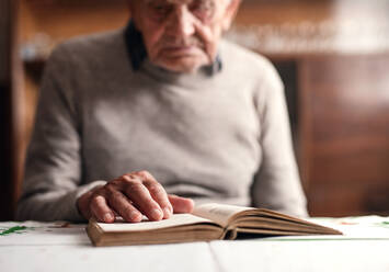 A portrait of elderly man sitting at the table indoors at home, resting and reading bible. - HPIF05872