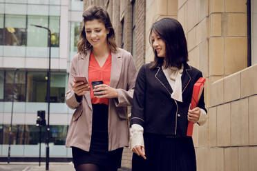 Smiling businesswoman with colleague using smart phone at footpath - PWF00560