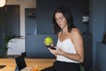 Junge Frau mit Smartphone und Apfel in der Hand zu Hause - PNAF04822