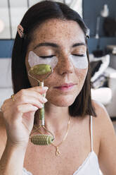 Woman with under eye patches massaging using jade stone roller on face at home - PNAF04847