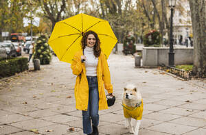 Glückliche Frau, die einen gelben Regenschirm hält und mit ihrem Hund auf einem Fußweg spazieren geht - JCCMF08854