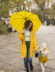 Glückliche schöne Frau, die einen Regenschirm hält und mit einem Hund auf einem Fußweg spazieren geht - JCCMF08858