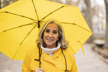 Ältere Frau mit gelbem Regenschirm auf dem Fußweg - JCCMF08863