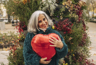 Glückliche ältere Frau mit rotem herzförmigen Ballon vor Pflanzen - JCCMF08883