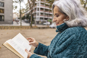 Lächelnde ältere Frau, die am Fußweg ein Buch liest - JCCMF08891