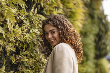 Smiling young woman standing in front of plants - JCCMF08905