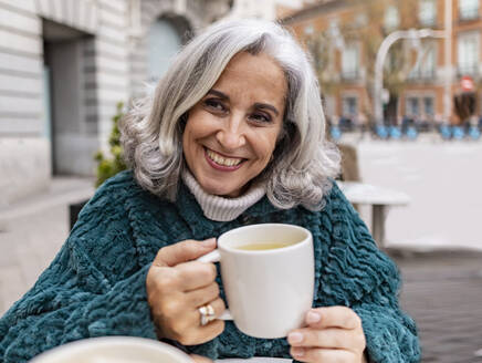 Happy senior woman with coffee cup sitting in sidewalk cafe - JCCMF08928