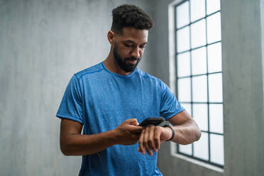 A happy young African American sportsman standing indoors at gym, using smartphone and smartwatch. - HPIF05909