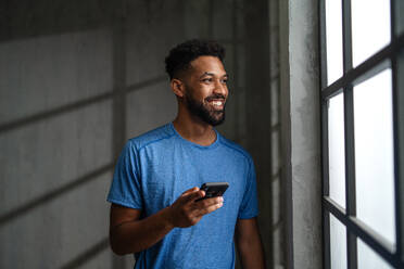 A happy young African American sportsman standing indoors at gym, using smartphone. - HPIF05912