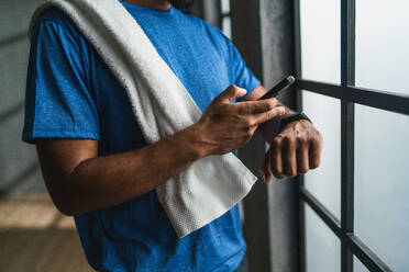 An unrecognizable young African American sportsman standing indoors at gym, using smartphone and smartwatch. - HPIF05918