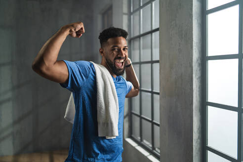 Ein glücklicher junger afroamerikanischer Sportler, der in einem Fitnessstudio steht, in die Kamera schaut und Muskeln zeigt - HPIF05930