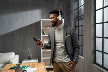 Ein glücklicher junger afrikanischer amerikanischer Geschäftsmann mit Smartphone und Headset arbeitet drinnen im Büro - HPIF05932