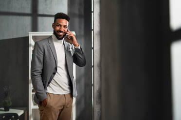 Ein glücklicher junger afroamerikanischer Geschäftsmann mit martphone drinnen im Büro, der in die Kamera schaut. - HPIF05940