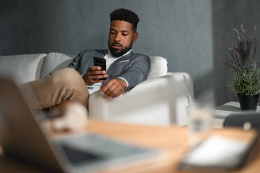Ein junger afroamerikanischer Geschäftsmann mit Smartphone sitzt auf dem Sofa in einem Büro - HPIF05941