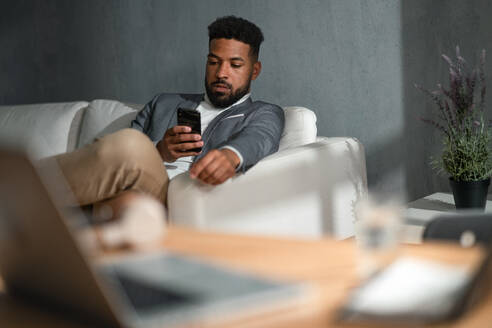 A young african american businessman with smartphone sitting on sofa indoors in office - HPIF05941