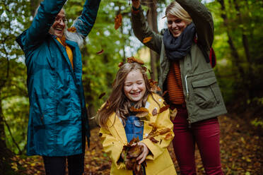 Ein glückliches kleines Mädchen mit Mutter und Großmutter, die sich bei einem Herbstspaziergang im Wald mit Blättern vergnügen - HPIF06021