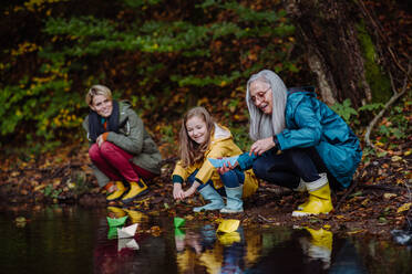 A small girl with mother and grandmother playing with paper boats in lake outoors in nature. - HPIF06033