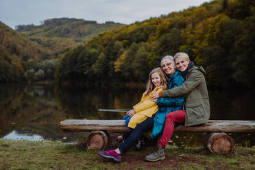 Ein kleines Mädchen mit Mutter und Großmutter sitzt auf einer Bank und schaut in die Kamera, draußen am See. - HPIF06036