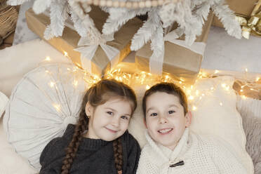 Lächelndes Mädchen mit Bruder bei Weihnachtsbeleuchtung und Geschenken zu Hause liegend - ONAF00368