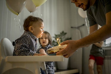 Vater mit Sohn, der Kerzen auf dem Kuchen ausbläst und seinen Geburtstag mit der Familie zu Hause feiert - ANAF00842