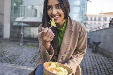 Glückliche junge Frau beim Mittagessen vor einem Gebäude sitzend - JCCMF08943