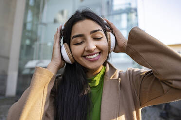 Glückliche Frau mit Kopfhörern, die vor einem Gebäude Musik hört - JCCMF08945
