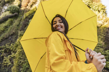 Smiling young woman holding yellow umbrella in front of vertical garden - JCCMF08964