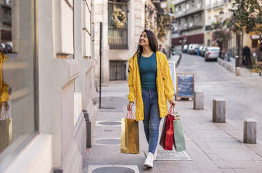 Happy young woman walking with shopping bags on footpath - JCCMF08969