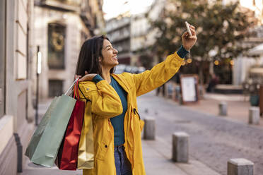 Happy woman with shopping bags taking selfie over smart phone on footpath - JCCMF08971