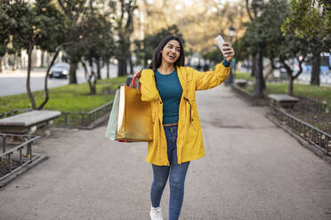 Glückliche Frau mit Einkaufstüten, die ein Selfie mit ihrem Handy auf dem Gehweg macht - JCCMF08972