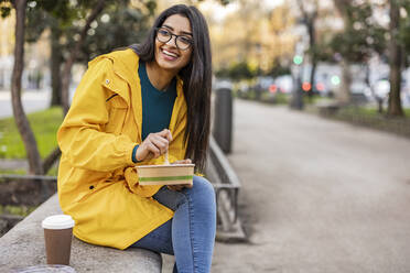 Happy young woman eating snack on bench in park - JCCMF08975