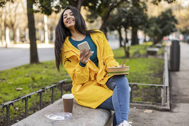 Happy young woman with bowl and smart phone sitting on seat - JCCMF08977