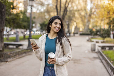 Nachdenklich lächelnde junge Frau mit Smartphone im Park stehend - JCCMF08980