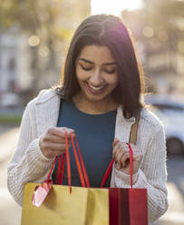 Smiling young woman looking in shopping bags on sunny day - JCCMF08986