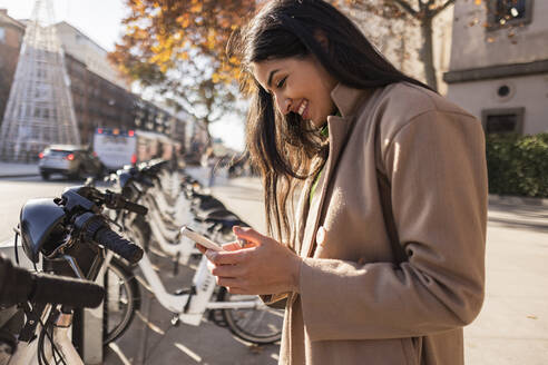 Glückliche junge Frau, die ein Smartphone benutzt und an einer Fahrradabstellanlage steht - JCCMF09012