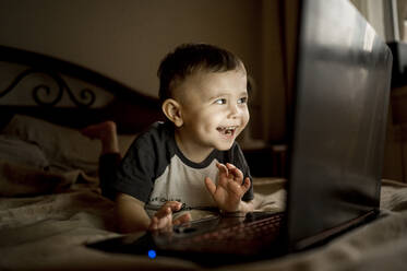 Glücklicher Junge mit Laptop auf dem Bett liegend zu Hause - ANAF00855