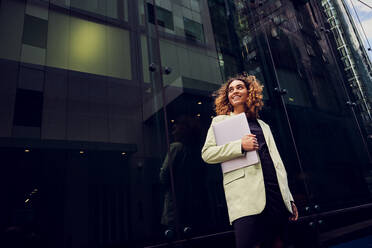 Smiling businesswoman with laptop standing near glass building - PWF00662