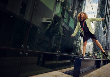 Verspielte Geschäftsfrau balanciert auf einer Bank in der Nähe eines Bürogebäudes - PWF00667