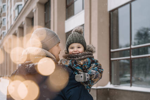 Vater trägt seinen Sohn vor einem Gebäude im Winter - ANAF00878