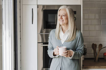 Glückliche reife Frau mit Kaffeetasse in der Küche stehend - EBBF07642