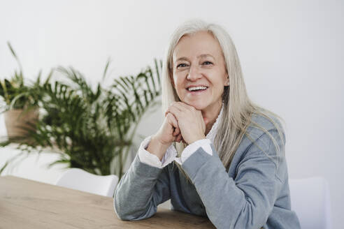 Happy mature woman with hand on chin sitting at table - EBBF07647